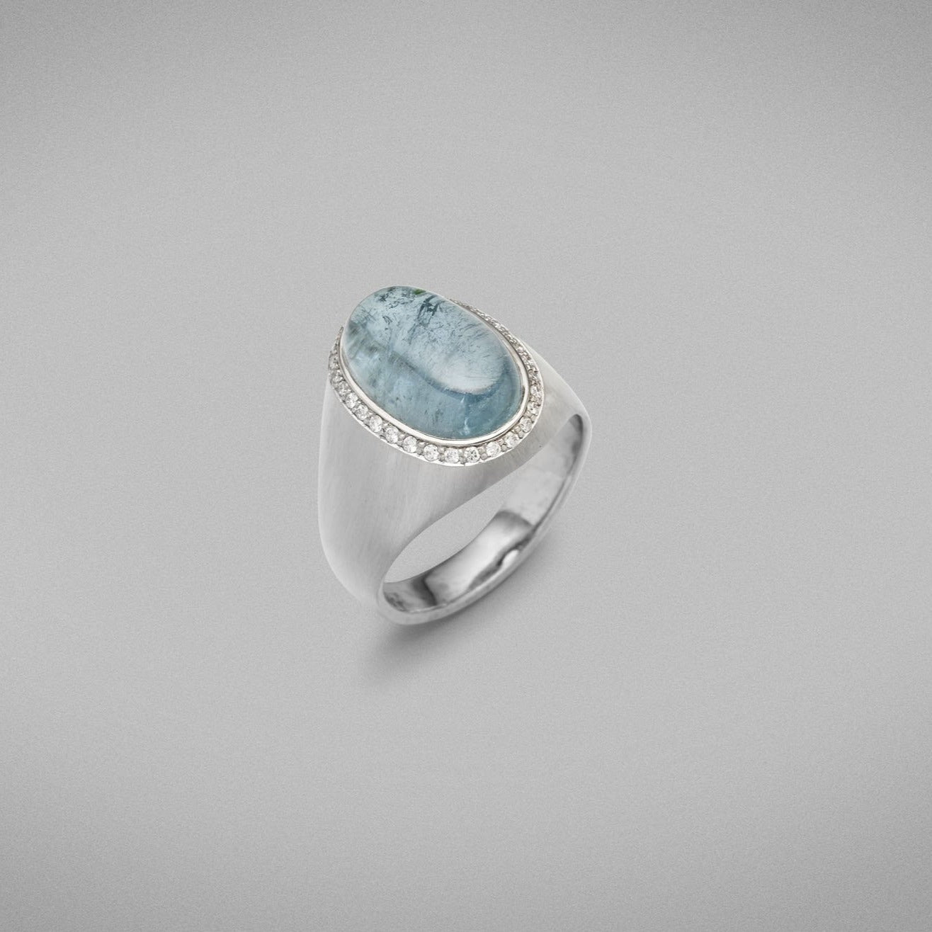 A BUNDA "Corvus" ring mounted in all 18 carat white gold and bezel set with a single oval cabochon pale blue Tourmaline, surrounded with a line of thread set diamonds. 18ct white gold setting has elegant brushed gold finish.  The Tourmaline weighs 6.50 carats.  Characteristics of diamonds: 31 = 0.15ct, F colour, VS clarity.  Weight of Ring: 9.40 grams.